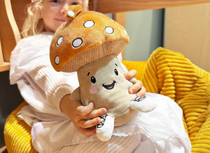 Ein Kind sitzt entspannt auf einem großen gelben Kissen und hält den Plüschpilz Felix vor sich. Felix trägt einen großen braun-orangefarbenen Pilzhut mit weißen Punkten, hat ein fröhliches Gesicht mit rosa Bäckchen und trägt kleine, gestickte Sneaker an den Füßen. Das Kind lächelt und präsentiert Felix in einer gemütlichen Spielsituation.