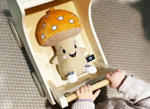 Ein kleines Kind schiebt einen hellen Holzwagen über einen Teppich. Darin sitzt Felix, der beigefarbene Plüschpilz mit großem orangebraunem Hut und weißen Punkten. Felix lächelt freundlich, seine rosa Bäckchen und die kleinen gestickten Sneaker an den Füßen sind gut sichtbar.