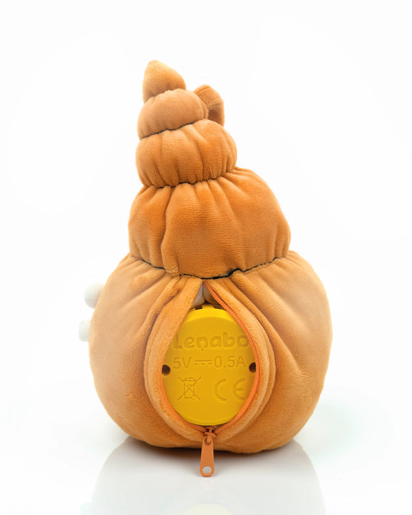 Der muschelförmige Lenabo Charakter Mia ist von hinten zu sehen. Das orangefarbene Plüschtier ist über einen Reißverschluss geöffnet, sodass der gelbe Lenabo Smart Speaker sichtbar wird. Auf dem Lautsprecher stehen technische Angaben wie ‚5V — 0.5A‘ und CE-Symbole.