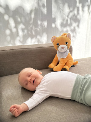 Ein schlafendes Baby liegt entspannt auf einem hellgrauen Sofa, während Tali, der gelb-orange Puma-Plüschcharakter mit grauem Halstuch, aufrecht daneben sitzt. Weiches, gefiltertes Licht fällt durch einen Vorhang und wirft ruhige Schattenmuster an die Wand, wodurch eine friedliche und geborgene Atmosphäre entsteht.