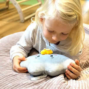 Ein Kleinkind liegt gemütlich auf einem weichen Kissen und hält Lenabie Lina (Wolke) sanft mit beiden Händen; sie schaut Lina neugierig an und lauscht dabei einer Hörspielwiedergabe von Lina, welche über die Lenabo App bequem von Eltern eingestellt und gesteuert wird. Das Kind blickt ruhig und aufmerksam auf Lina herab, wodurch eine warme, beruhigende Stimmung entsteht.