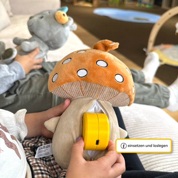 Ein Kind hält den Pilz-Plüschcharakter mit geöffnetem Reißverschluss auf dem Schoß und setzt den gelben Lenabo Smart Speaker in das Innenfach ein. Im Hintergrund sitzt ein anderes Kind und hält den grauen Bären-Plüschcharakter Aaron. Rechts unten befindet sich ein gelb umrahmtes Infofeld mit dem Text ‚einsetzen und loslegen‘.