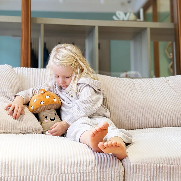 Ein blondes Kind sitzt gemütlich auf einem hellen, gerippten Sofa und hält den Lenabo Charakter Felix im Arm. Felix ist ein pilzförmiges Plüschtier mit braunem Hut und weißen Punkten sowie einem lächelnden Gesicht. Das Kind trägt einen hellen, gerippten Einteiler.