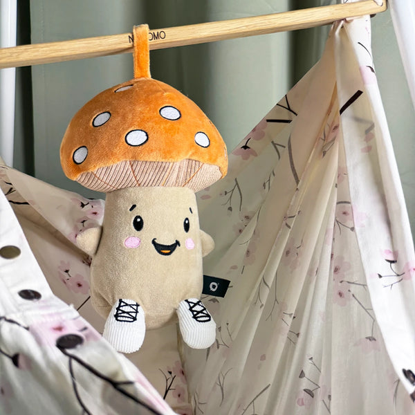 Der Lenabo Charakter Felix, ein pilzförmiges Plüschtier mit braunem Hut und weißen Punkten, hängt mithilfe einer Stoffschlaufe an einer Holzstange. Darunter befindet sich eine helle Baby-Hängematte mit feinem Blütenmuster in Rosa- und Beigetönen.