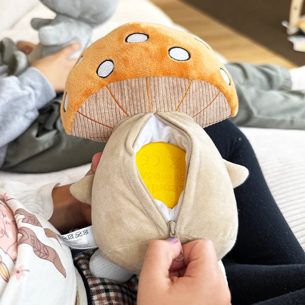 Ein Kind hält den Pilz-Plüschcharakter Felix auf dem Schoß und öffnet den Reißverschluss auf dessen Rückseite. Im Inneren ist der gelbe Lenabo Smart Speaker sichtbar. Im Hintergrund sitzt ein weiteres Kind und spielt mit dem grauen Bären-Plüschcharakter Aaron. Die Szene wirkt alltäglich und zeigt, wie der Speaker in die Figuren eingesetzt wird.