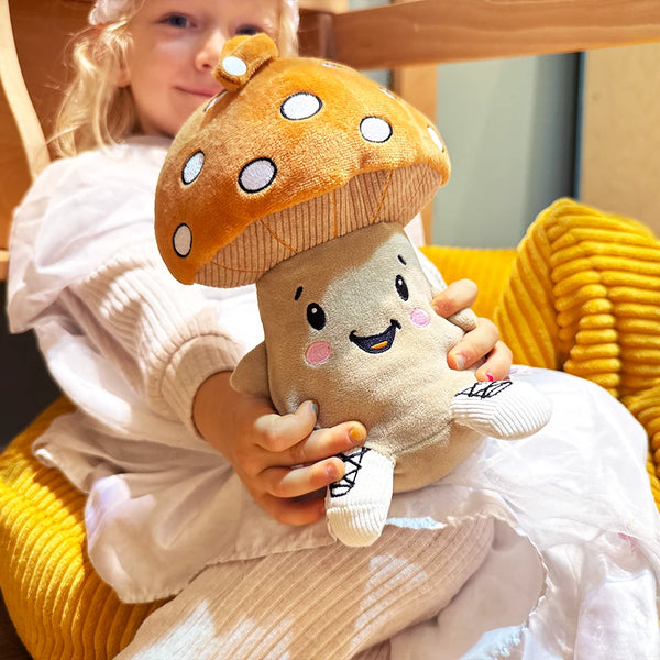 Ein Kind sitzt auf einem gelben Kissen und hält das Plüschtier Felix von Lenabo vor sich. Felix ist ein beige-brauner Pilz mit großem orangem Hut mit weißen Punkten, gesticktem freundlichen Gesicht und kleinen weißen Schuhen. Das Kind hält ihn mit beiden Händen nach vorne gerichtet.