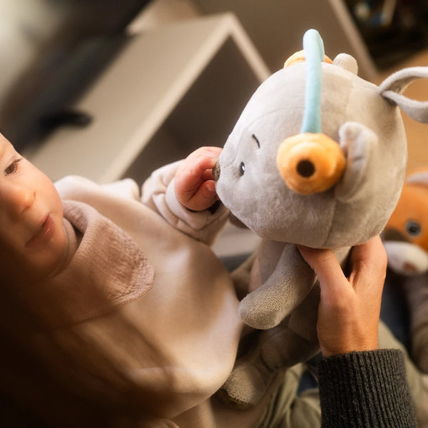 Ein kleines Kind sitzt auf dem Schoß einer erwachsenen Person und berührt sanft das Gesicht des grauen Bären Aaron, einem Lenabo-Kuscheltier mit blauem Bügel und gelben Ohrwärmern. Die erwachsene Person hält Aaron dabei fest.