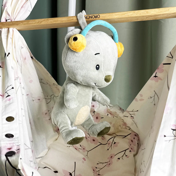 Das graue Lenabo-Kuscheltier Aaron mit türkisfarbenem Bügel und gelben Ohrwärmern hängt an einer Holzstange einer NONOMO-Federwiege. Die Wiege besteht aus einem hellen Stoff mit zarten Kirschblütenmustern.“