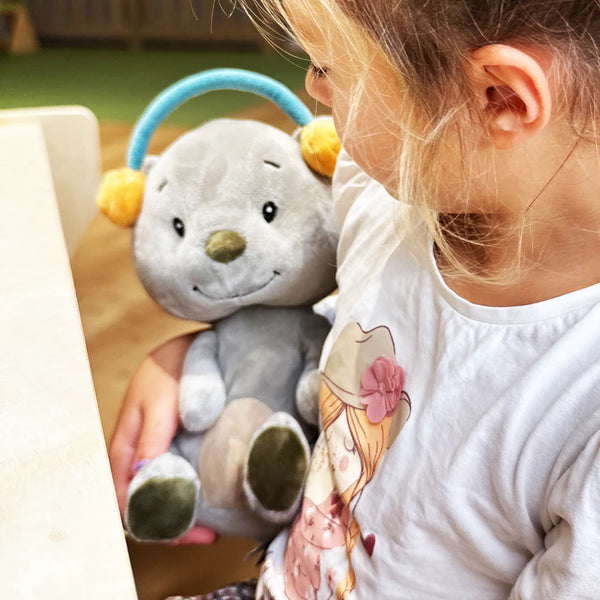 Ein kleines Kind sitzt an einem Tisch und hält das graue Kuscheltier Aaron von Lenabo im Arm. Aaron trägt einen türkisfarbenen Bügel mit gelben Ohrwärmern. Das Kind schaut lächelnd auf das Kuscheltier.