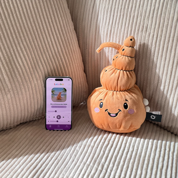 „Der Lenabo Charakter Mia, eine orangefarbene muschelförmige Stofffigur mit fröhlichem Gesicht, sitzt auf einem hellen, gerippten Sofa. Daneben steht ein Smartphone mit geöffneter Lenabo App, auf dem die Gute-Nacht-Geschichte ‚Die schimmernde Welle‘ angezeigt wird, inklusive Wiedergabesteuerung.
