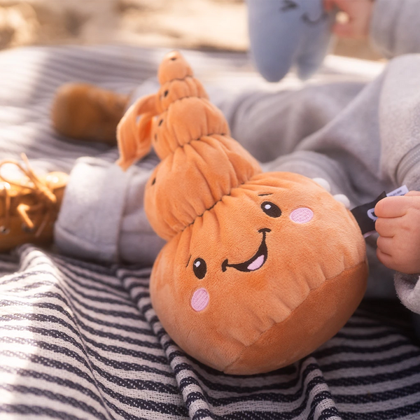 Der Lenabo Charakter Mia liegt auf einer dunkelblau-weiß gestreiften Decke und wird von einem Kind gehalten, das graue Kleidung und braune Schuhe trägt. Mia ist muschelförmig, orangefarben und zeigt ein fröhliches Gesicht mit rosa Wangen. Im Hintergrund ist ein weiterer Lenabo Charakter leicht verschwommen zu sehen.