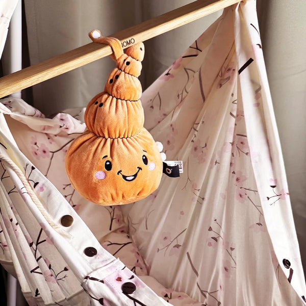 Der Lenabo Charakter Mia hängt mit ihrer Schlaufe an der Holzstange einer Baby-Hängematte. Mia ist eine orangefarbene, muschelförmige Stofffigur mit fröhlichem Gesicht und rosa Wangen. Die Hängematte besteht aus einem hellen Stoff mit feinem Blütenmuster in Rosatönen.