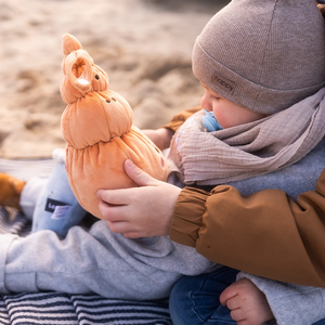 Ein kleines Kind sitzt auf einer gestreiften Decke am Strand und hält den Lenabo Charakter Mia, eine orangefarbene muschelförmige Stofffigur mit freundlichem Gesicht. Das Kind trägt graue Kleidung, eine braune Jacke, eine Mütze und einen Schal.