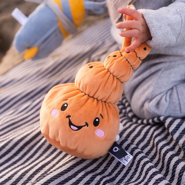 Ein Kind sitzt auf einer dunkelblau-weiß gestreiften Decke und hält den Lenabo Charakter Mia, eine orangefarbene muschelförmige Stofffigur mit fröhlichem Gesicht und rosa Wangen, an ihrer Schlaufe fest. Im Hintergrund liegt der blaue Lenabo Charakter Nalu leicht unscharf.