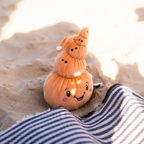 Der Lenabo Charakter Mia, eine orangefarbene muschelförmige Stofffigur mit fröhlichem Gesicht und rosa Wangen, sitzt auf hellem Sand. Sonnenlicht wirft leichte Schatten auf ihre Oberfläche. Im Vordergrund liegt eine dunkelblau-weiß gestreifte Decke.