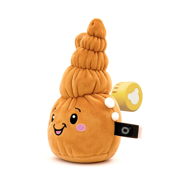 Der muschelförmige Lenabo Charakter Mia ist in seitlicher Ansicht zu sehen. Das orangefarbene Plüschtier mit mehreren rundlich gestapelten Segmenten hat ein freundliches Gesicht mit rosa Wangen. Auf der Rückseite ist der gelbe Lenabo Smart Speaker sichtbar und teilweise aus der Öffnung herausgezogen.