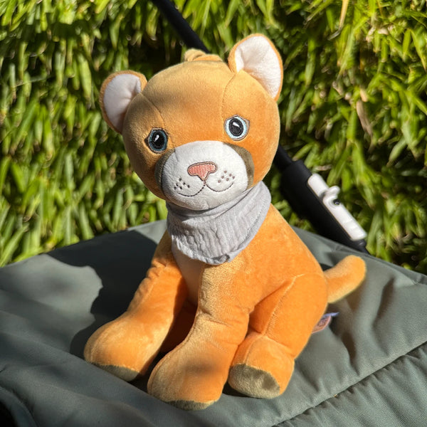 Der gelbe Plüschpuma Tali sitzt auf dem Sitz eines Kinderwagens. Hinter ihm ist dichter grüner Bambus zu sehen, und warmes Sonnenlicht fällt auf sein Gesicht.