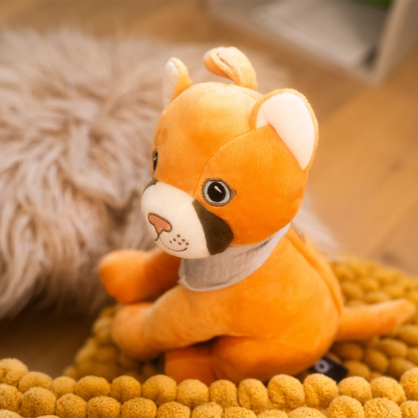 Der gelbe Plüschpuma Tali sitzt leicht seitlich gedreht auf einem weichen, gelb strukturierten Kissen. Er trägt sein graues Halstuch und blickt mit großen, freundlichen Augen nach vorn. Im Hintergrund liegt ein flauschiges, beiges Kissen auf einem hellen Holzboden. Die Szene wirkt warm und gemütlich.