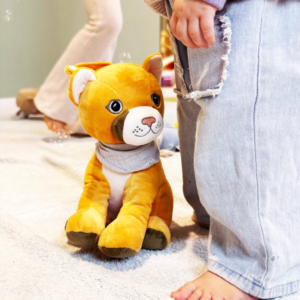 Plüschpuma Tali sitzt aufrecht auf einem weichen, hellen Teppich. Sein graues Halstuch liegt locker um den Hals. Direkt neben ihm steht ein Kind, von dem nur Beine und Hände sichtbar sind. Im Hintergrund sind weitere spielende Kinder zu sehen. Das Bild zeigt eine warme, kindliche Spielumgebung.