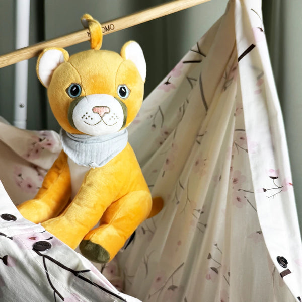 Der gelbe Plüschpuma Tali sitzt in einer hellen Baby-Hängematte mit zartem Kirschblütenmuster. Er trägt ein graues Halstuch und hängt mit einer kleinen Stoffschlaufe an einer Holzstange über der Hängematte. Der Hintergrund ist weich und neutral gehalten. Die Szene wirkt ruhig und liebevoll arrangiert.
