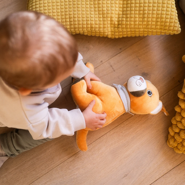 Ein kleines Kind sitzt auf einem Holzboden und hält den gelben Plüschpuma Tali mit beiden Händen fest. Tali liegt seitlich vor dem Kind und trägt sein graues Halstuch. Um das Kind herum liegen gelbe, strukturierte Kissen. Die Szene wirkt warm, ruhig und spielerisch.