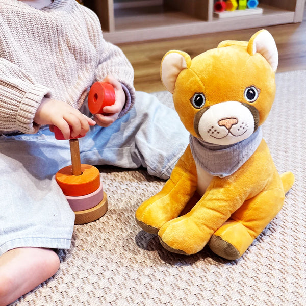 Ein kleines Kind sitzt auf einem hellen Teppichboden und spielt mit einem Holzsteckspiel aus runden, bunten Ringen. Direkt daneben sitzt Tali, der gelbe Plüschpuma mit grauem Halstuch, aufrecht und dem Kind zugewandt. Die Szene wirkt warm, ruhig und zeigt ein kindgerechtes Spielumfeld.