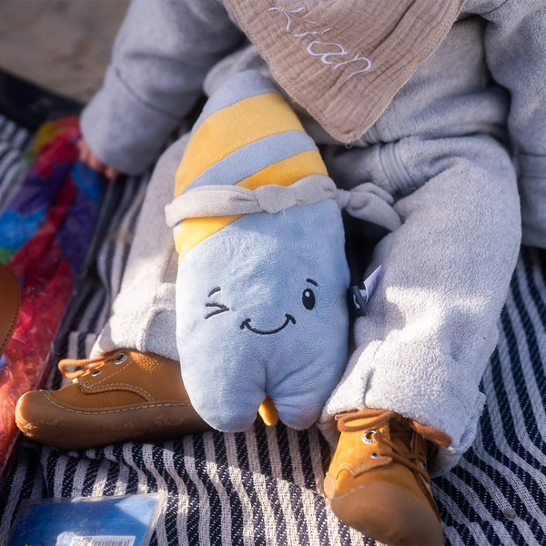 Ein Kind sitzt in grauer Kleidung auf einer gestreiften Decke und hält den Lenabo Plüschcharakter Nalu auf dem Schoß. Nalu ist blau-gelb gestreift, trägt ein graues Halstuch und lächelt mit einem zwinkernden Gesicht.