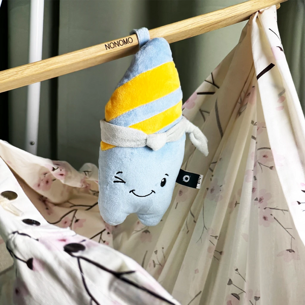 Der Lenabo Plüschcharakter Nalu hängt mit einer Schlaufe an einer Holzstange der NONOMO Babyhängematte. Nalu ist blau mit zwei gelben diagonalen Streifen und einem grauen Halstuch. Die Hängematte besteht aus einem hellen Stoff mit zarten floralen Mustern.