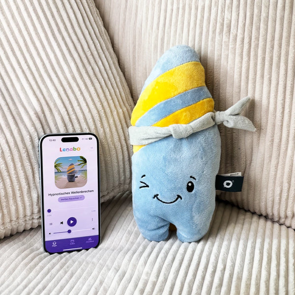 Der Lenabo Charakter Nalu steht auf einem hellen Stoffuntergrund. Rechts neben ihm ist ein Smartphone platziert, auf dem die Lenabo App geöffnet ist. Die App zeigt ein Nalu-Audioprogramm mit dem Titel ‚Hypnotisches Wellenbrechen‘ und Bedienelementen für die Wiedergabe.