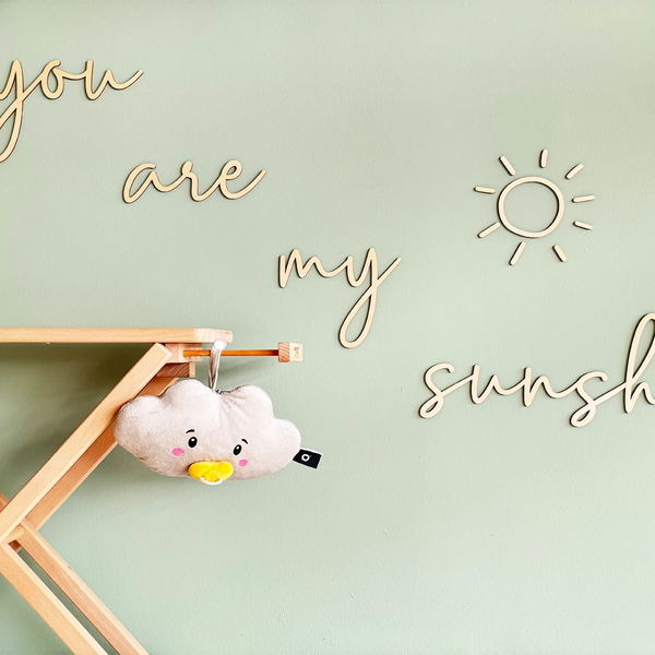 Der wolkenförmige Lenabo Charakter Lina hängt mithilfe einer Stoffschlaufe an einem hellen Holzgestell. Der Hintergrund zeigt eine mintgrüne Wand mit dekorativen Holzschriftzügen, darunter die Worte ‚you are my sunshine‘ und eine stilisierte Sonne.